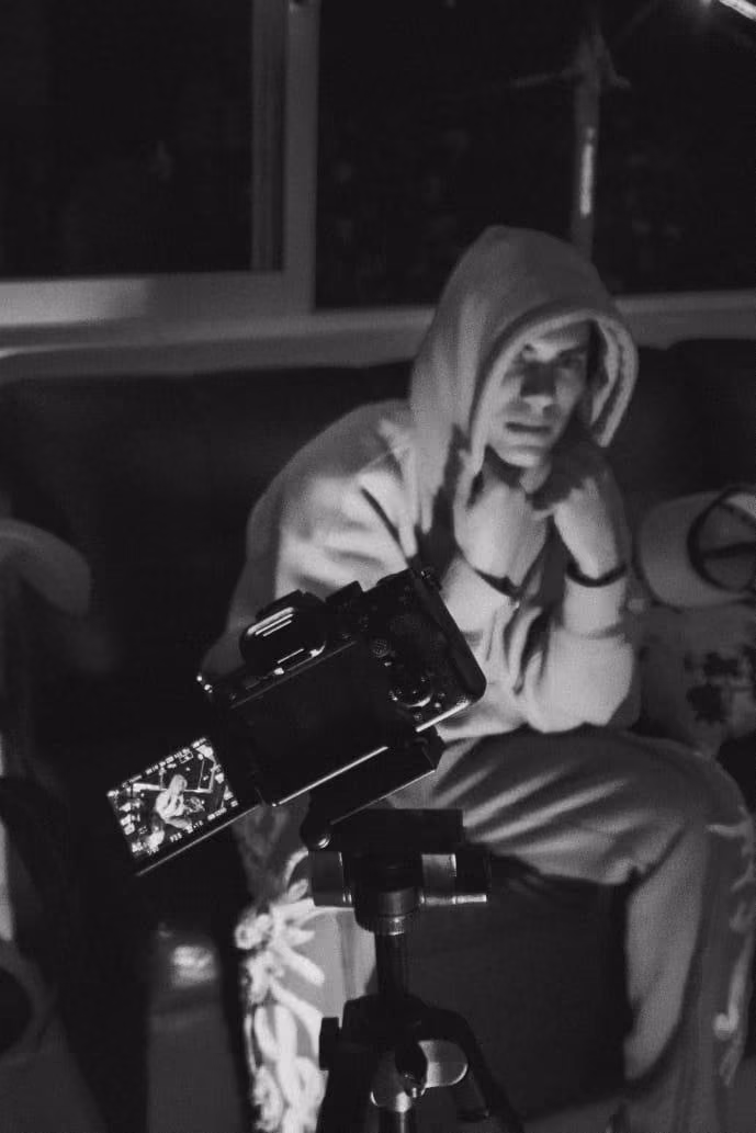 Black-and-white photo of an artist in a hoodie seated behind a camera on a tripod.