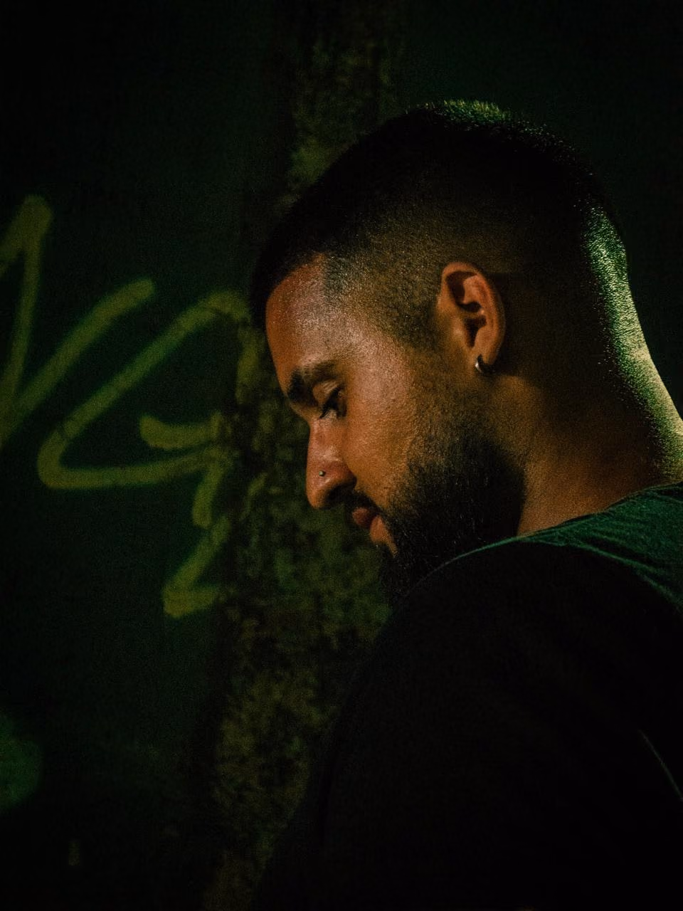 Profile portrait in green light against a wall with graffiti.