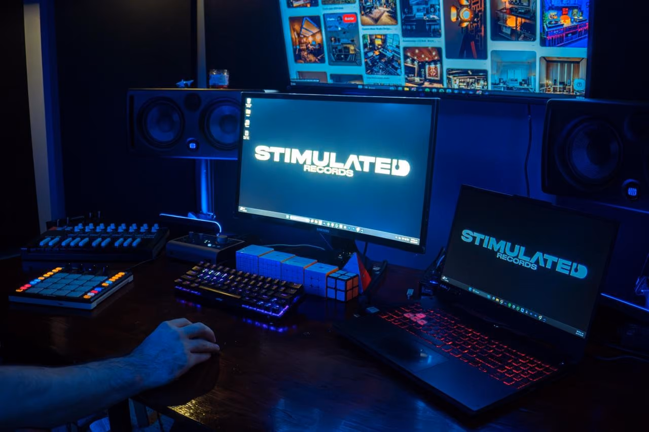 Studio desk in blue light with the Stimulated Records logo on a monitor and a laptop.